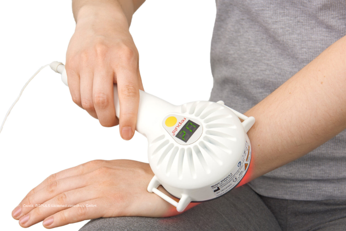 Zu sehen ist ein repuls Tiefenstrahler, der mit seinem kalten Rotlicht zur Schmerztherapie an der Hand zum Einsatz kommt.